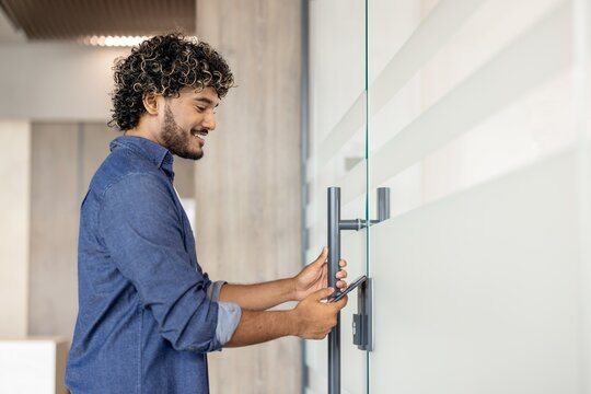 Smiling young Indian man opens the door to the office with an electronic key on his phone
