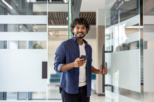 Portrait of a young Indian man holding a phone in his hand, entering the office, opening the door and looking at the camera