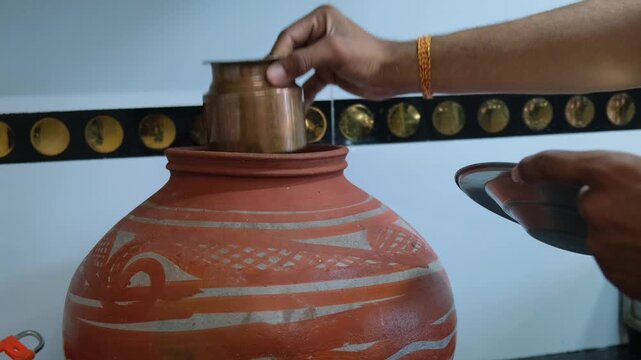 Pouring fresh water from a traditional earthen pot into a metal tumbler in an Indian kitchen