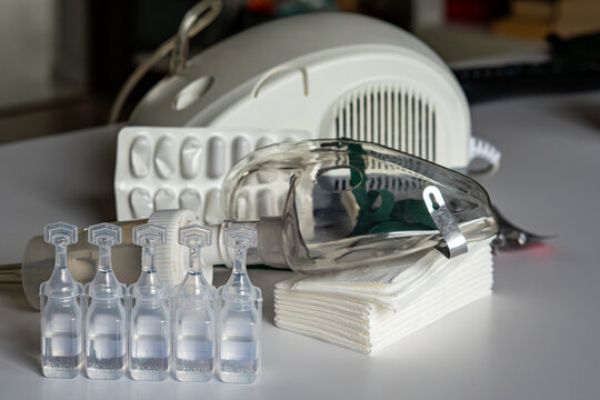 Medical nebulization set featuring a row of five saline solution ampoules in the foreground, a transparent inhalation mask, a stack of white tissues, and a white compressor inhaler in the background, 