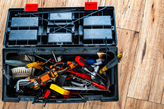 Open black plastic toolbox, top view looking inside the cluttered interior filled with assorted hand tools like pliers, screwdrivers, brush, and zip ties, resting on a light wood plank floor, natural 