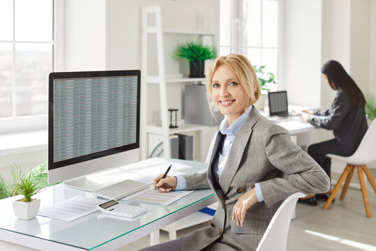 Cheerful confident businesswoman sitting at desk in office and working with financial data on large monitor. Smiling professional accountant looking at camera while analyzing spreadsheet on desktop.