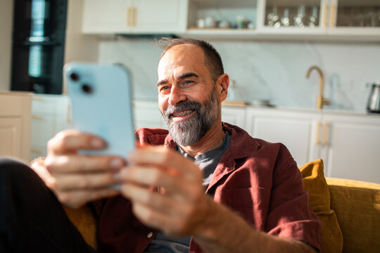 Smiling bearded man using smartphone on couch at home