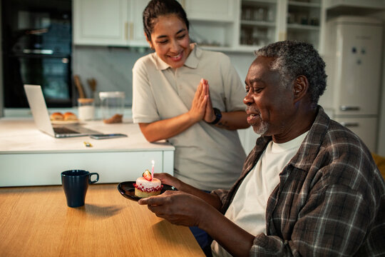 Caregiver celebrating senior man's birthday with cupcake and candle at home kitchen