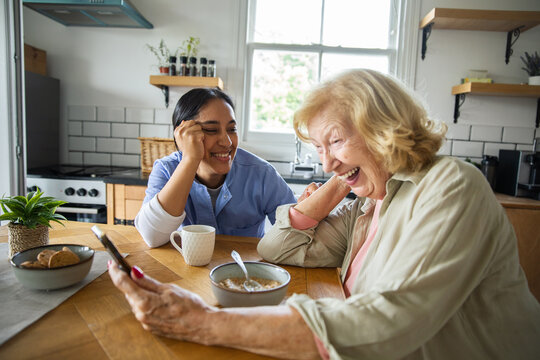 Caregiver and senior woman laughing with smartphone at home kitchen