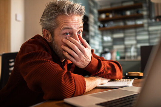 Tired middle-aged man rubbing eyes while working on laptop in a home kitchen