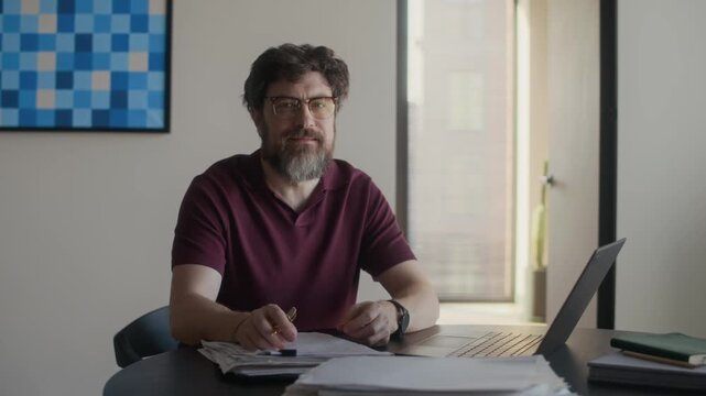 Medium portrait of middle aged Caucasian male writer sitting at table in living room, reading and editing novel draft then looking at camera