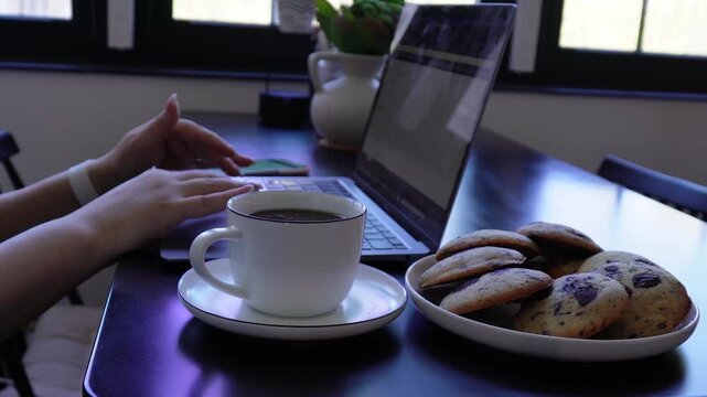 Female hands placing phone near laptop, typing, checking time, closing laptop and leaving desk with coffee and cookies remaining on table.