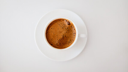 Top down view of a cup of frothy dark coffee in a white ceramic cup and saucer on a white background © İdil Toffolo