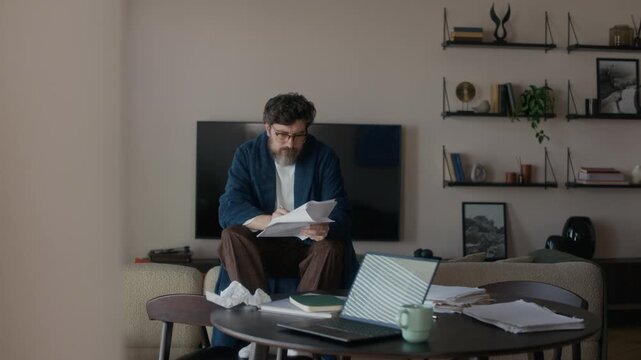 Middle aged Caucasian male scriptwriter wearing robe sitting on sofa in living room in his apartment editing printed draft