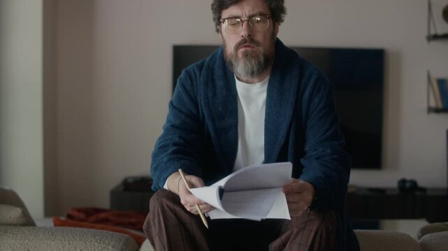 Mature Caucasian male author with beard on face wearing robe and eyeglasses sitting on sofa back, reading typescript, crumpling and throwing away badly written part