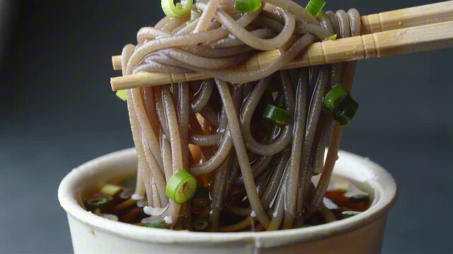 Cold soba noodles lifted by chopsticks from a cup of tsuyu sauce, showing wet buckwheat strands with a glossy sheen and floating green onion rings.