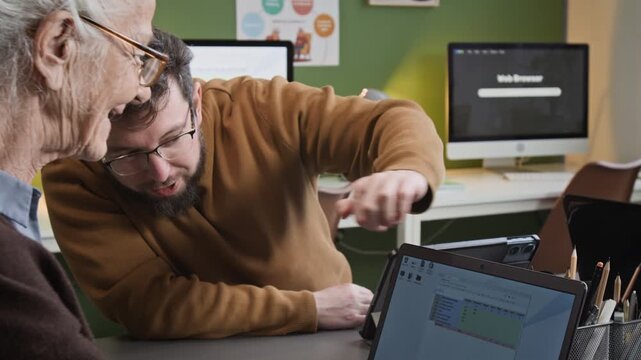 Tilt up shot of white haired senior woman using spreadsheets software during computer class for seniors with smiling male teacher helping and giving advice
