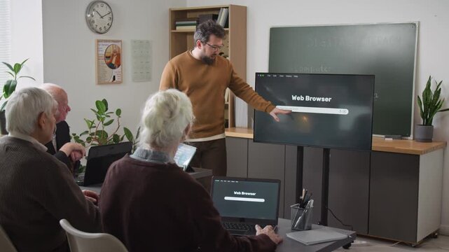 Medium shot of male teacher explaining internet basics to group of senior people in computer class and pointing at digital screen with web browser and search bar, copy space