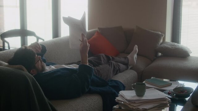 Middle aged Caucasian male writer wearing robe lying on sofa in living room, reading and correcting printed draft