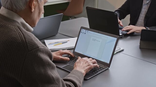 Over shoulder shot of senior man slowly typing on laptop keyboard and inputting text into digital document while attending computer literacy class, copy space