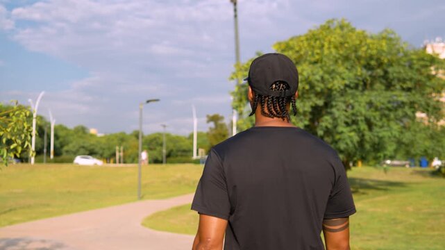 Young Black Latin man in sportswear looking back over shoulder while walking on path in sunny urban park daytime