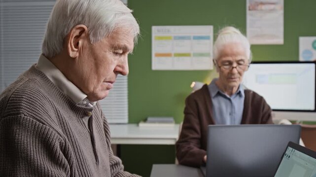 Tilt up side view shot of white haired senior man slowly typing text on laptop keyboard and looking at screen while sitting at desk in computer literacy class for seniors, copy space