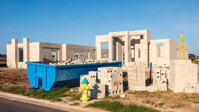 Street view of the construction site of one or two hurricane-resistant houses, with a fire hydrant, dumpster, and stacks of concrete blocks in foreground, in an upscale suburban development in Florida