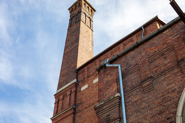 dramatic low angle view of a tall historic red brick tower rising above a matching brick building facade with decorative arched windows against a partly cloudy blue sky © Дмитрий Швец