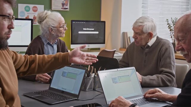 Side view shot of male teacher pointing at laptop screen while explaining tasks to senior man during computer literacy class in retirement home