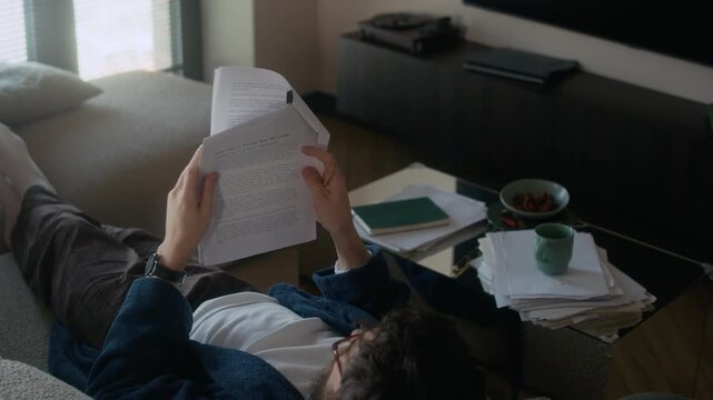 High angle of male writer reading novel typescript while lying on sofa in living room, tea mug, pile of papers and notebook visible on coffee table nearby