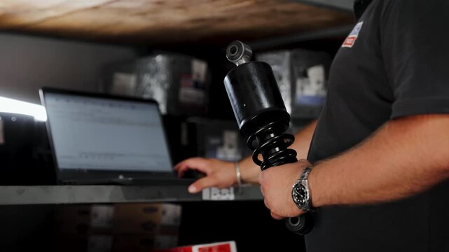 Mechanic checking car part information on laptop, holding a shock absorber for vehicle repair and maintenance