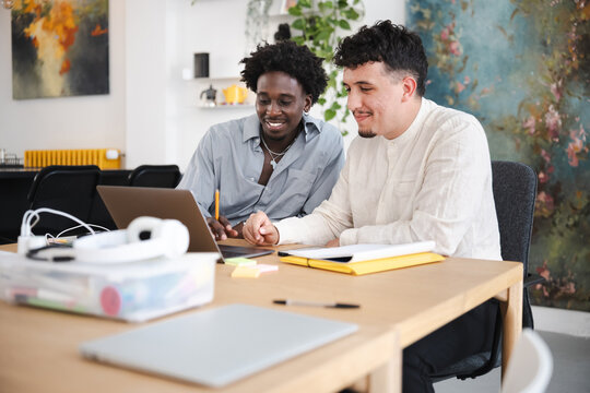 Two young men collaborate on a project at a desk, using a laptop and notebooks.