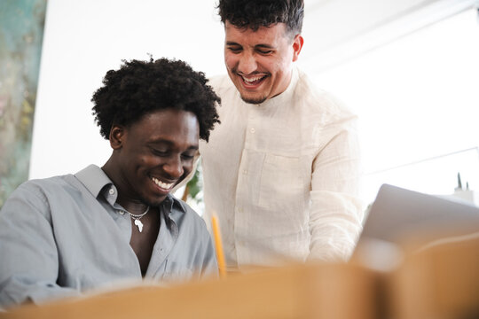 Two young men share a laugh while working together on a project, one looking down at a document and the other leaning in with a smile.