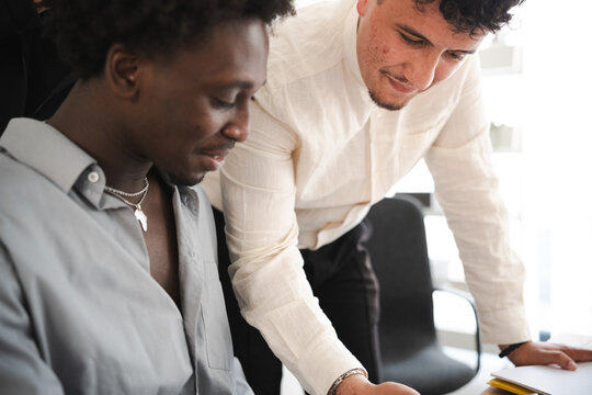 Two men collaborate on a project, one leaning over a desk to review documents with the other.