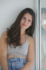 Fototapeta premium Young woman sitting by a window at home, smiling in soft natural daylight. Casual lifestyle portrait with relaxed mood and gentle reflection in glass.