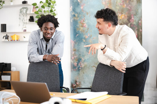 Two young men are engaged in a conversation while leaning on chairs in a modern office space.