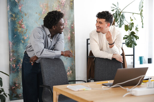 Two men in casual attire converse in a bright, modern office space, leaning on chairs near a large abstract painting and a desk with a laptop.