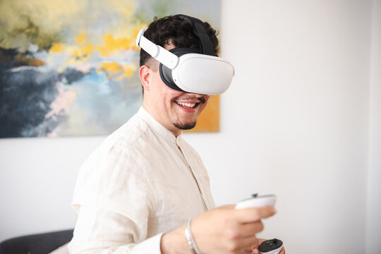 A man wearing a virtual reality headset and holding controllers smiles while experiencing immersive technology in a modern living room.