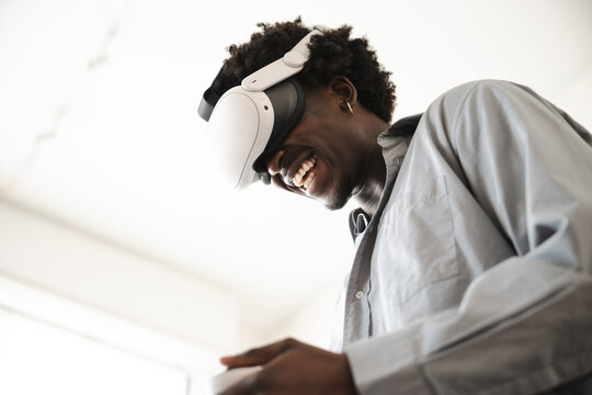 A young man with curly hair laughs while wearing a virtual reality headset. He is dressed in a light blue button-down shirt.