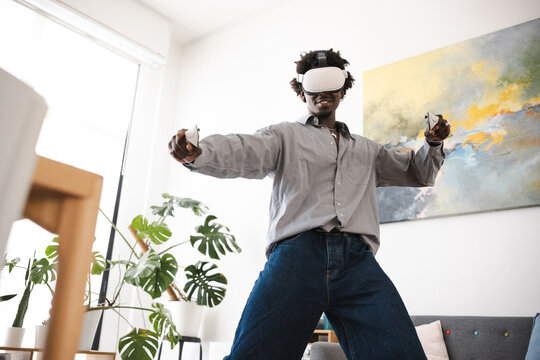 A young man wearing a virtual reality headset and holding controllers is immersed in a digital world, experiencing interactive entertainment at home.