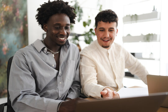 Two young men collaborate on a project, looking at a laptop screen with smiles and engaged expressions.