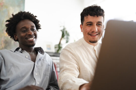 Two young men smile while looking at a laptop screen together, sharing a moment of collaboration or entertainment.