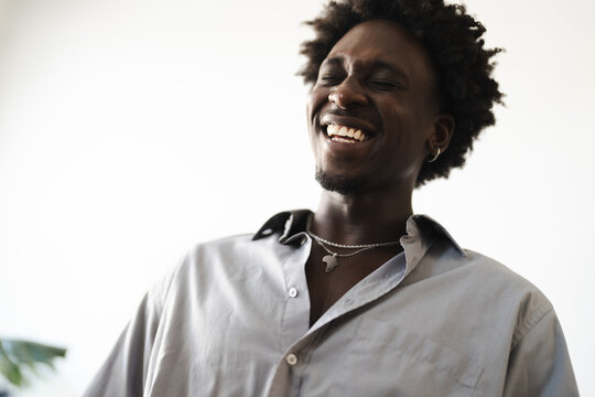 A young Black man with an afro laughs heartily, his eyes closed in joy. He wears a light grey button-down shirt and a silver necklace with a continent pendant.