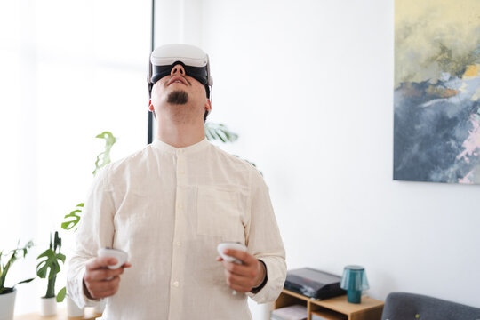 A man wearing a virtual reality headset and holding controllers looks up, immersed in a digital experience.