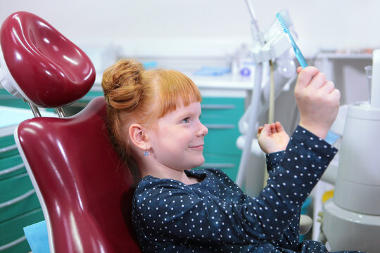 .Pediatric dentistry. Girl holds toothbrush in her hands. Cheerful child. Little girl with red hair is sitting in dental chair. Child is at dentist's office. Concept of oral hygiene and dental health