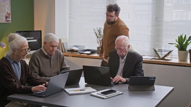 Group of senior people attending computer literacy classes and using laptops with male teacher overseeing lesson and giving instructions