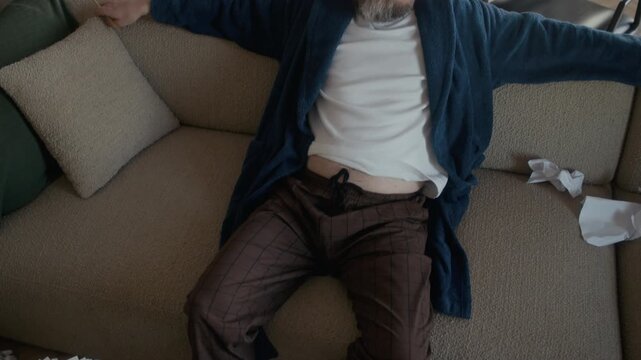 High angle view of exhausted middle aged Caucasian male writer procrastinating while sitting on sofa in messy living room