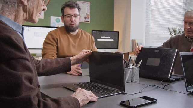 Medium shot of computer class for seniors starting with friendly teacher asking group of adult students to open laptops while sitting at desk in school