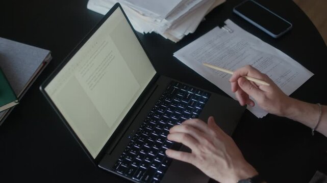 High angle view close up of unrecognizable male author writing novel on laptop, holding pencil in one hand and deleting bad part