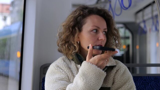 Caucasian woman with curly hair sitting by the window in a tram, holding a smartphone horizontally and recording a voice message while commuting through the city on a cloudy day