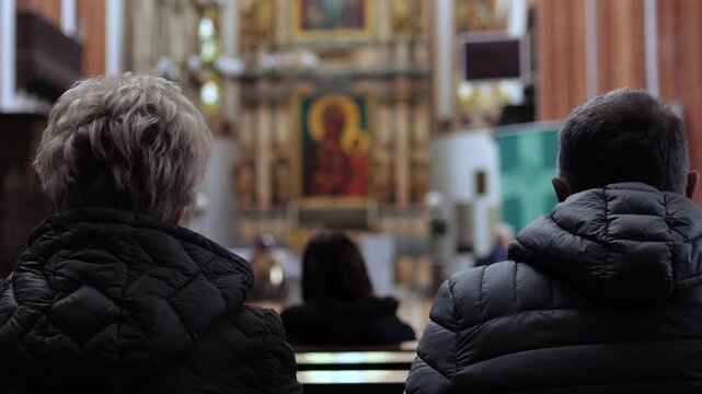Rear view of a congregation of faithful people sitting on pews during a religious sermon. An elderly woman and a man are praying silently in a beautiful and historic christian church