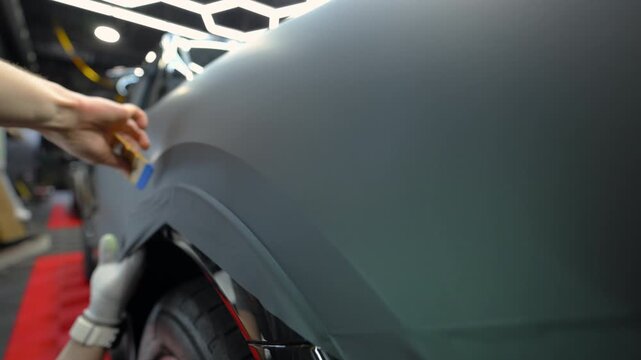 Close up of a technician applying matte vinyl wrap on a car body panel in a workshop. Automotive detailing process with protective film installation, smooth matte finish and precision work.