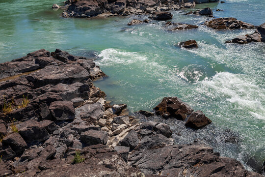 Sunlit turquoise river rapids flow around dark basalt rocks and a rugged shoreline, creating sparkling currents, whitewater splashes and natural textures ideal for outdoor, landscape and adventure