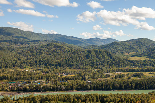 Katun river valley summer landscape photography. A peaceful rural village sits along a turquoise river at the foot of forested mountains under a bright blue sky with scattered clouds
